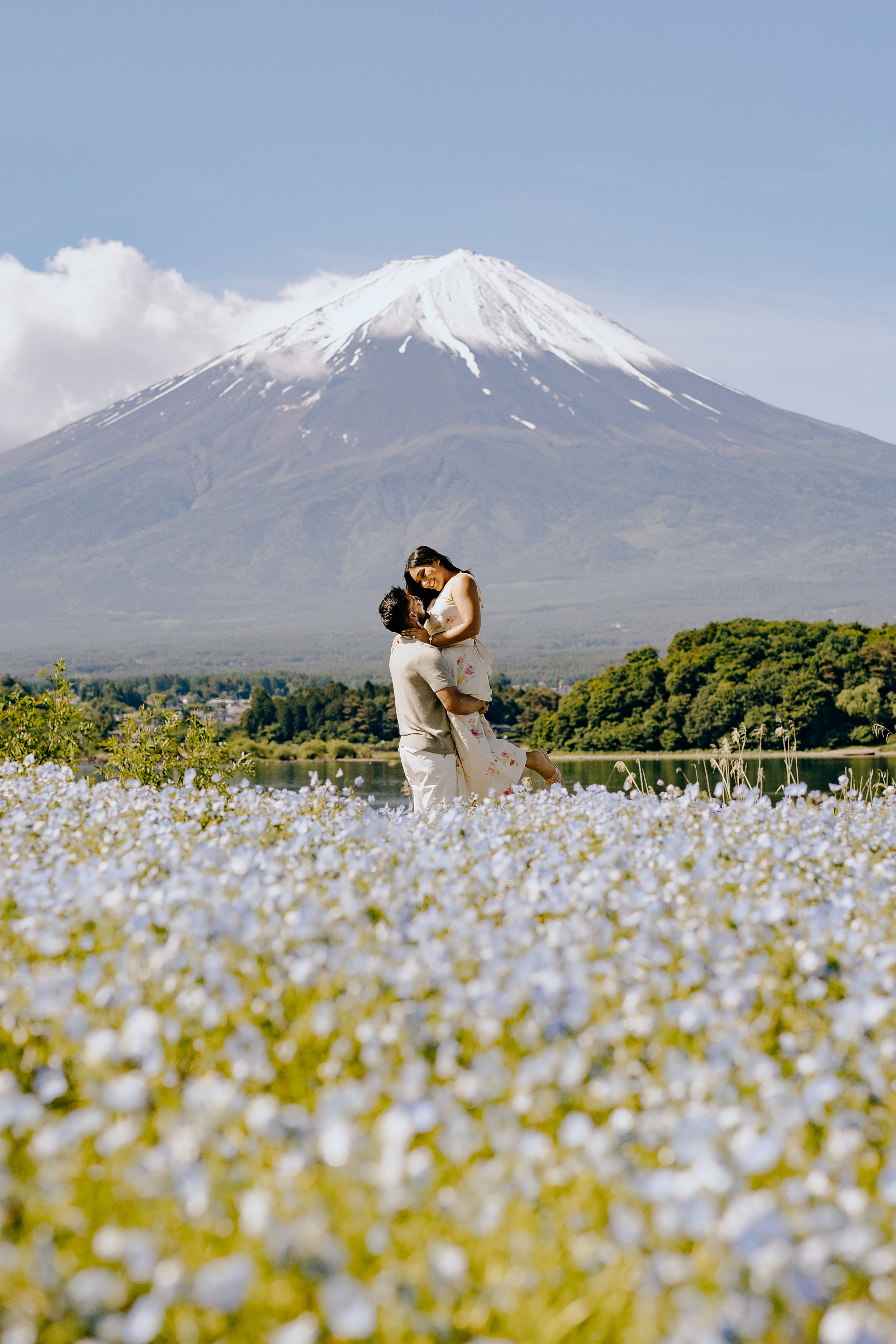 A couple embraces in a field of white flowers with majestic Mount Fuji rising in the background under blue skies.