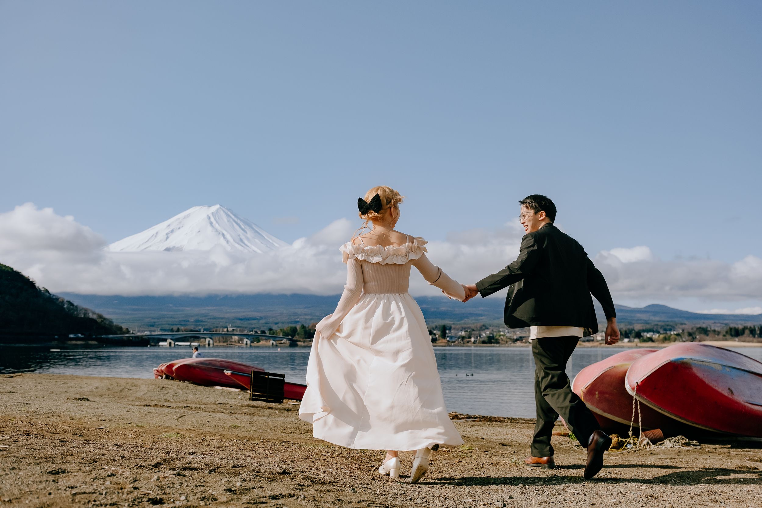 Couple in wedding attire holding hands on beach with Mount Fuji and red canoes in background on sunny day.
