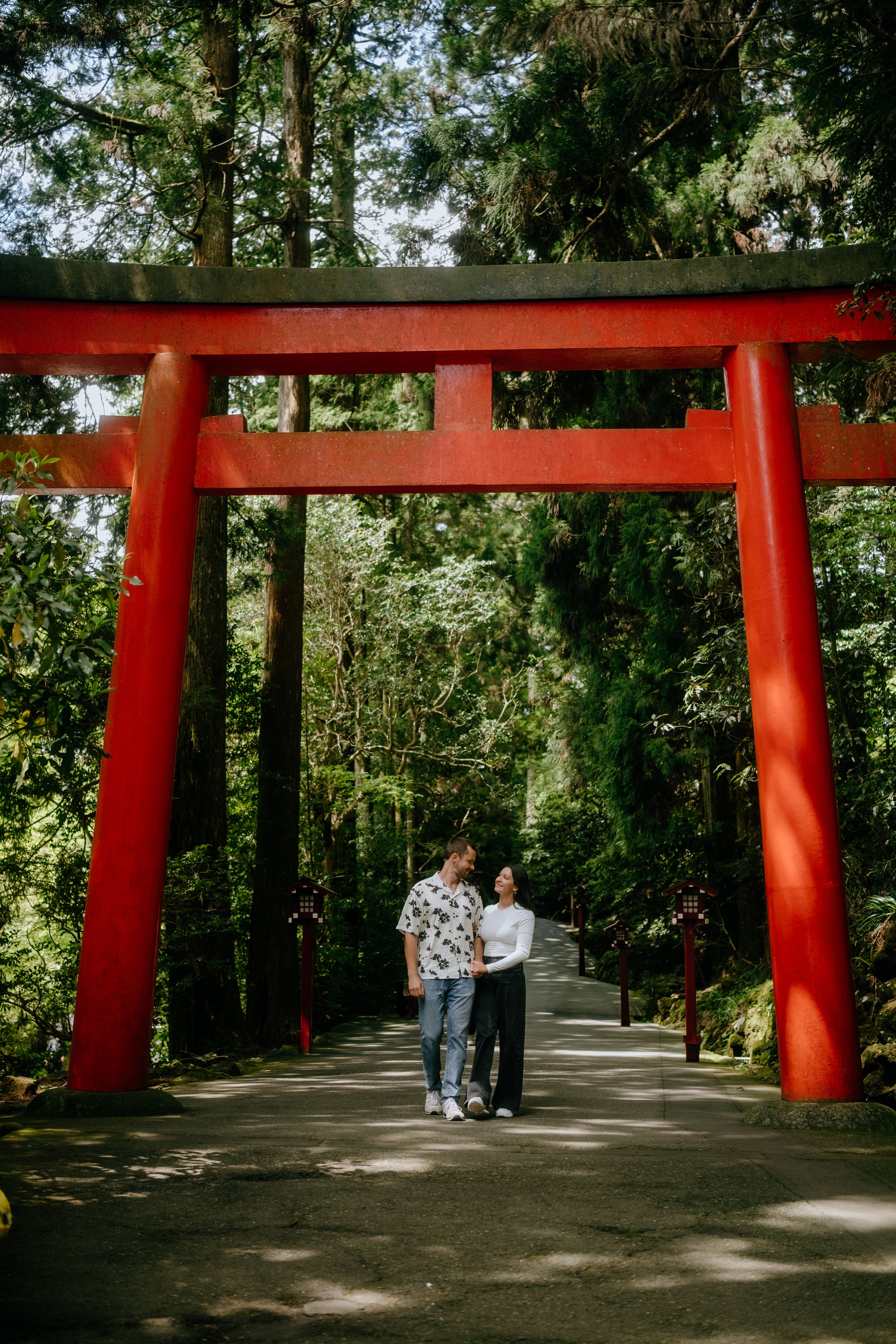 Couple holding hands under a red torii gate surrounded by lush green forest trees.