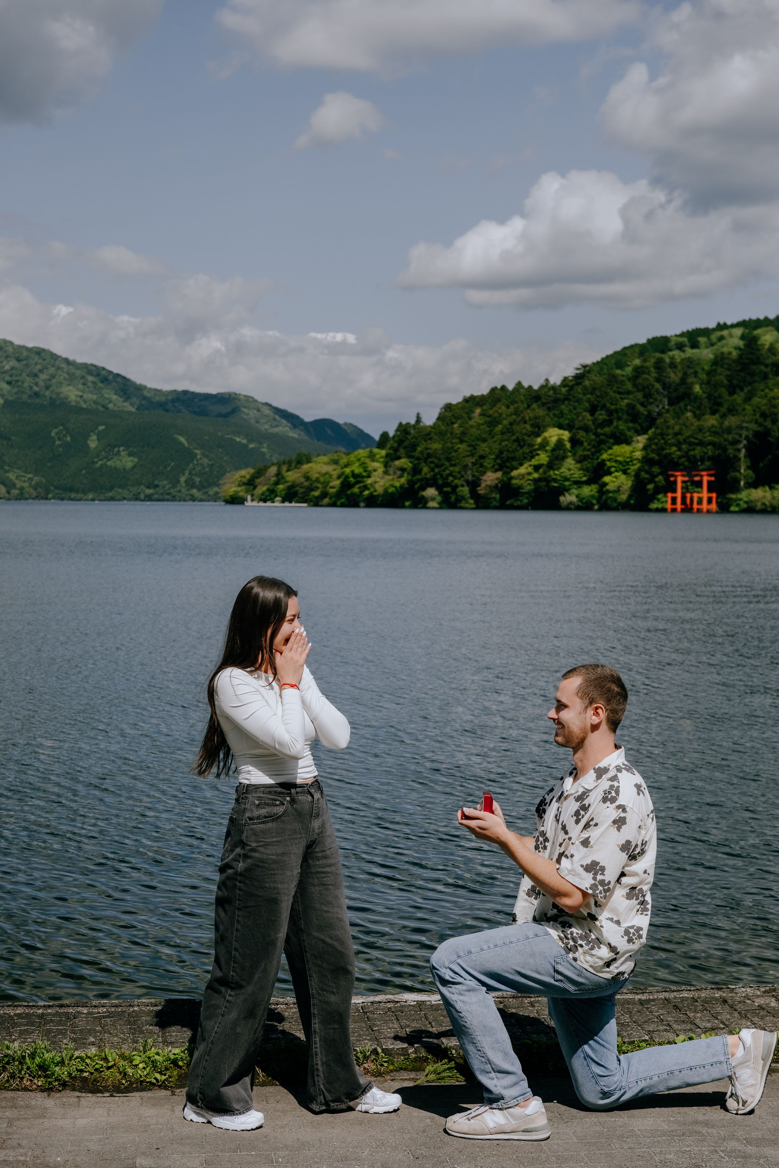 Man proposes on one knee to surprised woman by scenic lake with red torii gate in Japan.
