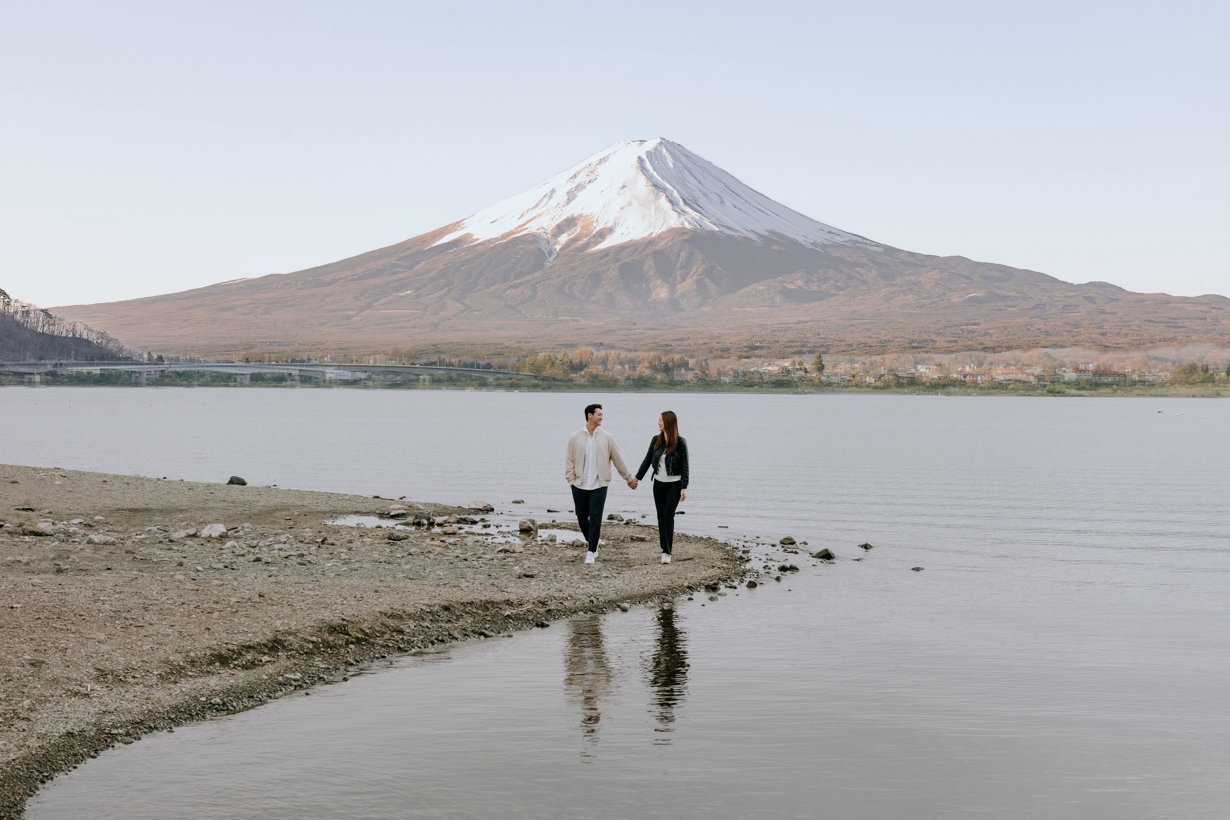 Couple holding hands walking along a lakeside shore with iconic snow-capped Mount Fuji in the background.