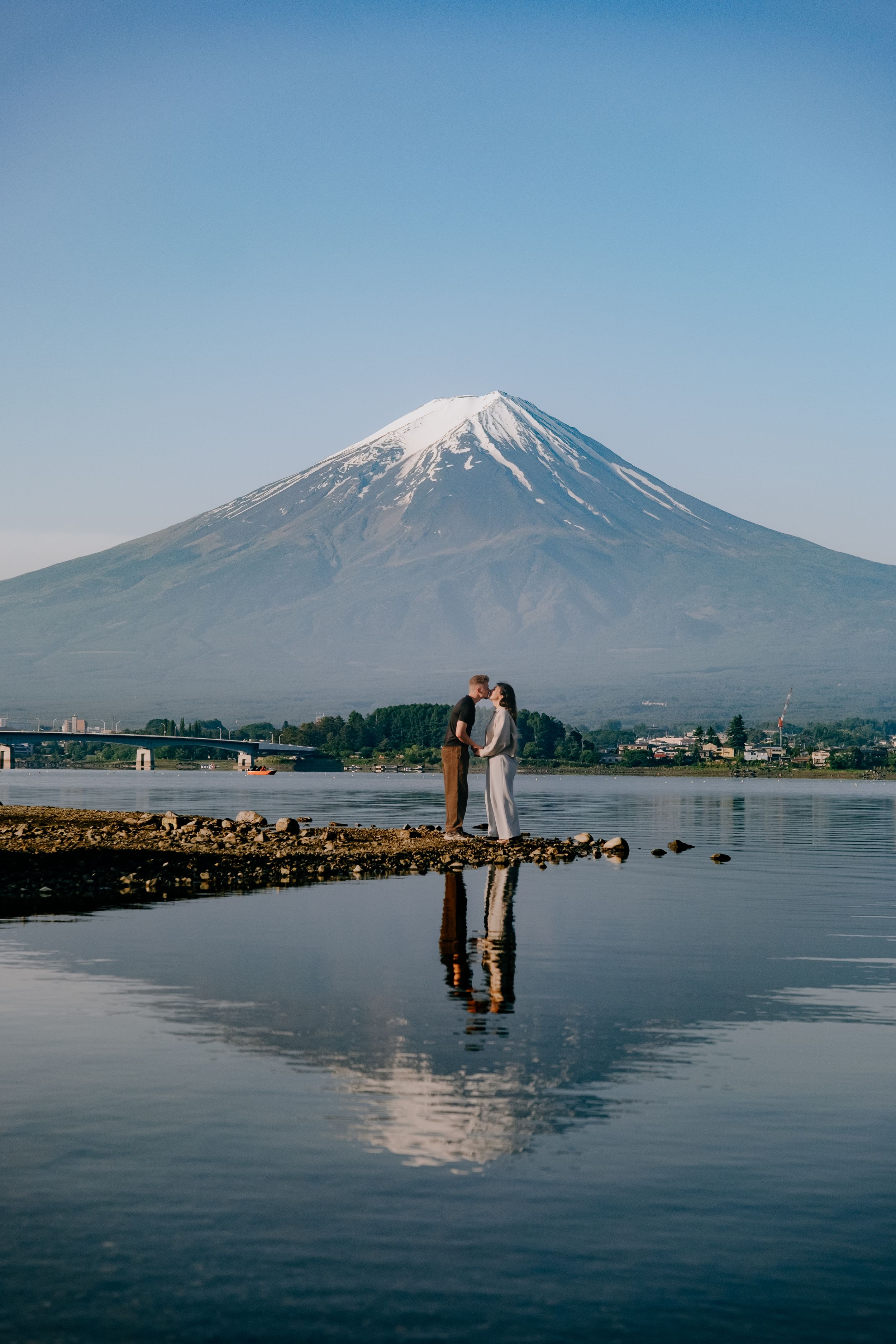 Couple kissing by Lake Kawaguchi with iconic snow-capped Mount Fuji reflected in calm water, Japan.