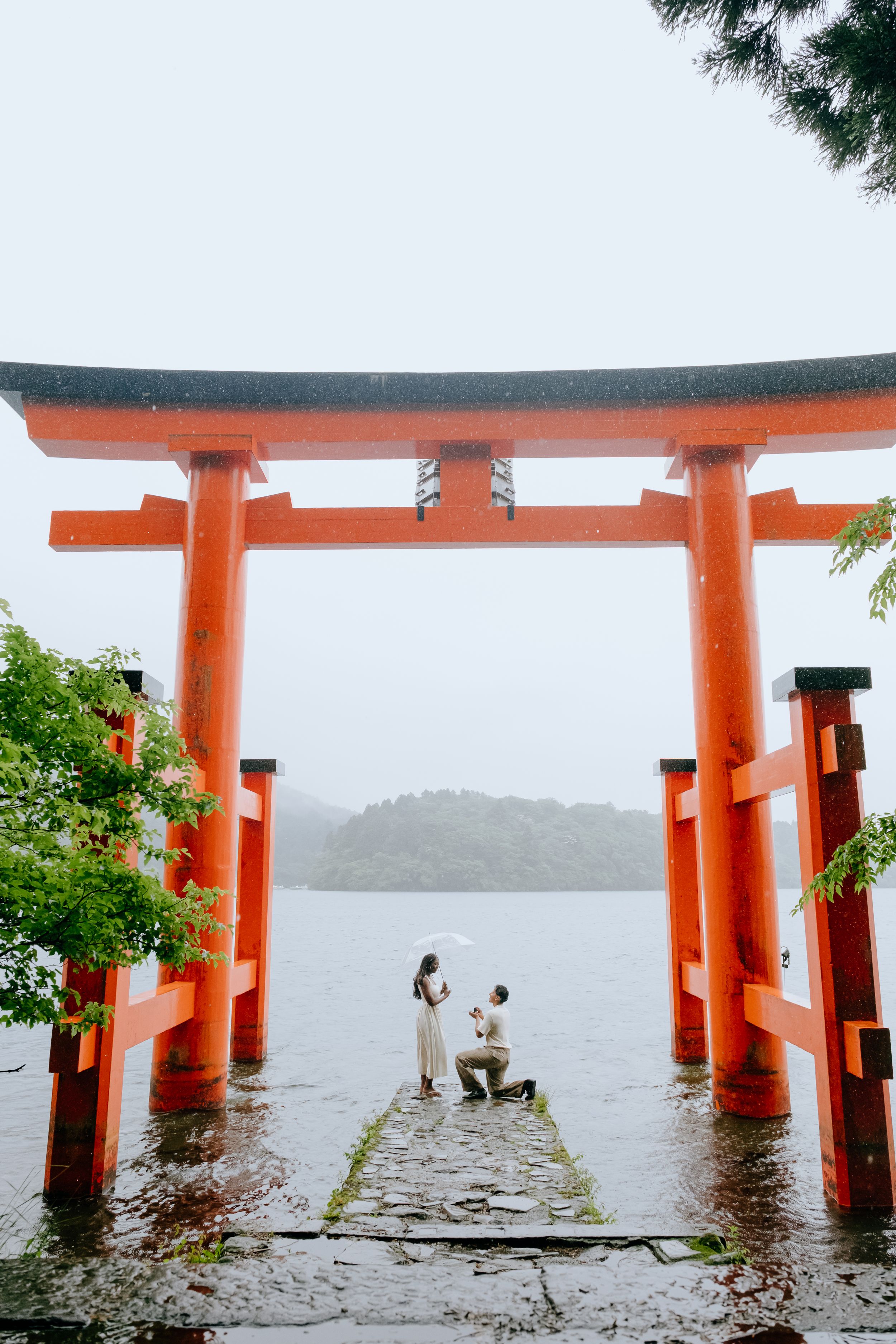 Couple's marriage proposal under a red torii gate at a Japanese shrine by a misty lake on a rainy day.