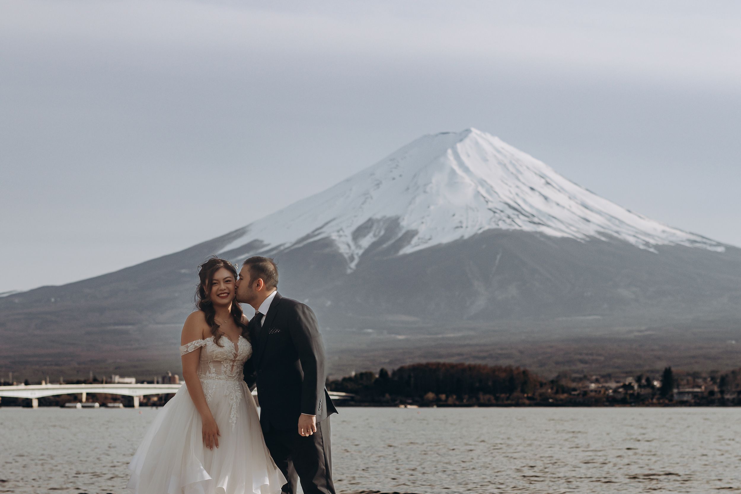 Wedding couple poses for romantic photos with Mount Fuji's snow-capped peak in the background at Lake Kawaguchiko.