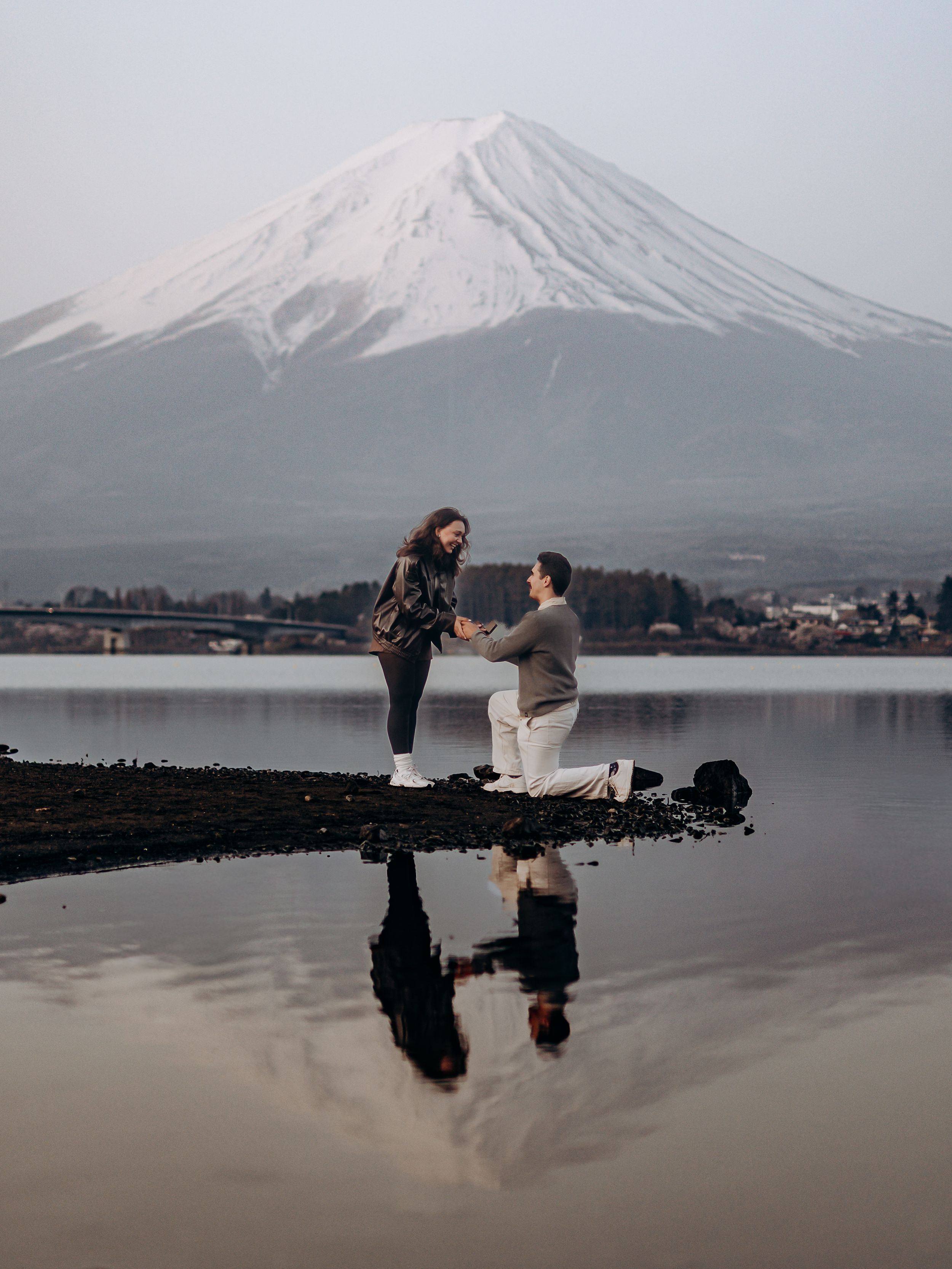 Mount Fuji's snowy peak reflects in Lake Kawaguchiko as couple shares romantic moment on jetty at sunset.