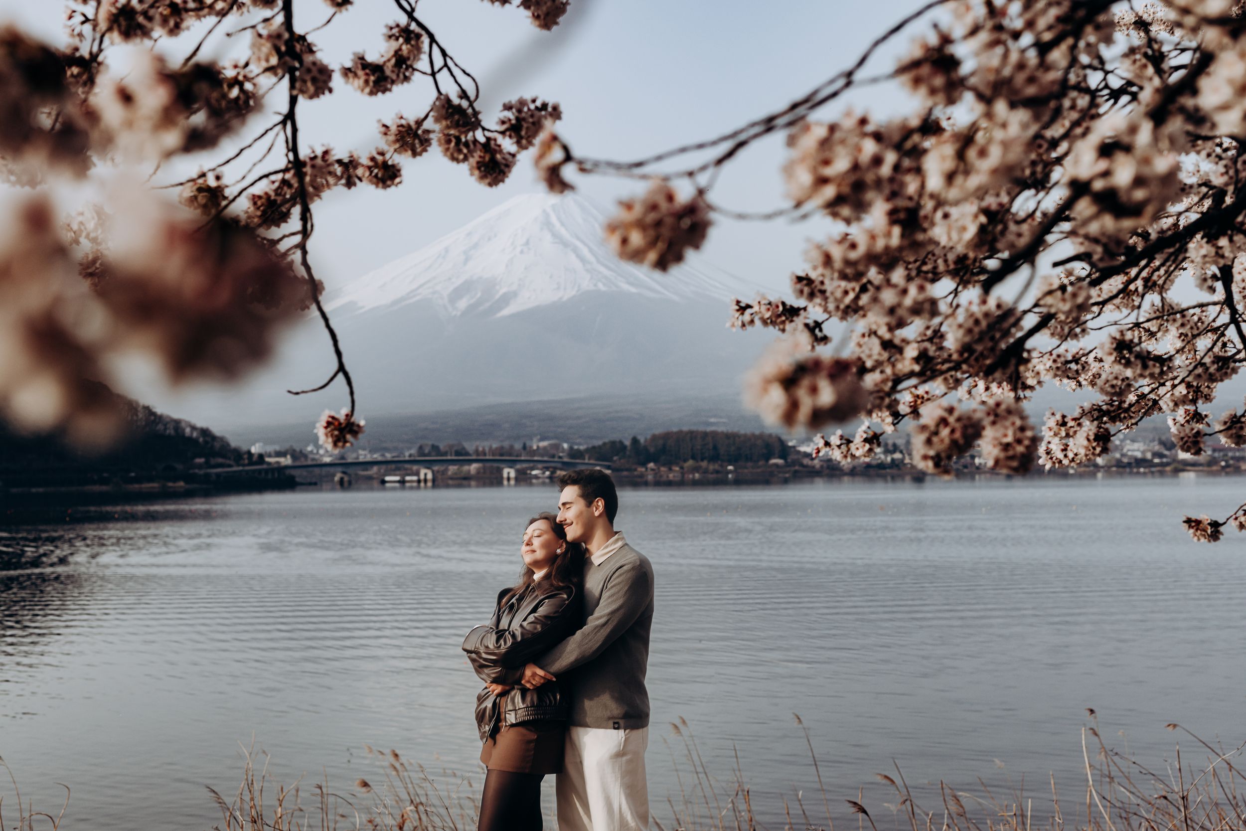 Couple embraces by serene lake with Mount Fuji and cherry blossoms framing scenic Japanese landscape.