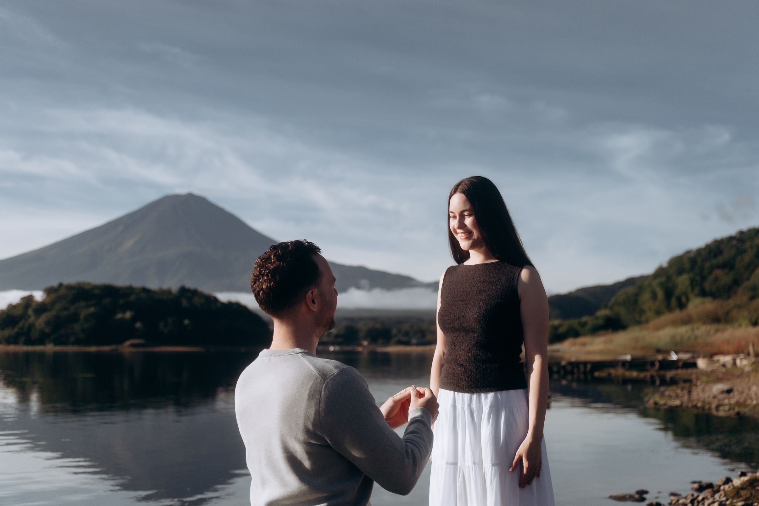 A couple in white outfits sits together on a dock overlooking a serene mountain lake at sunset.