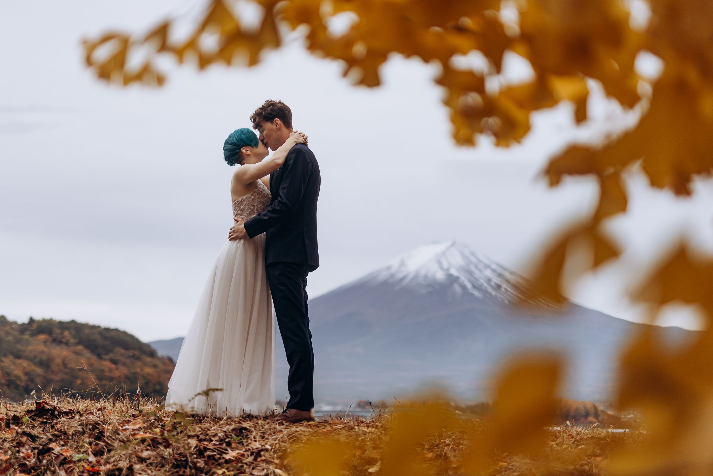 Couple in wedding attire embraces against autumn foliage and mountain backdrop during golden hour.