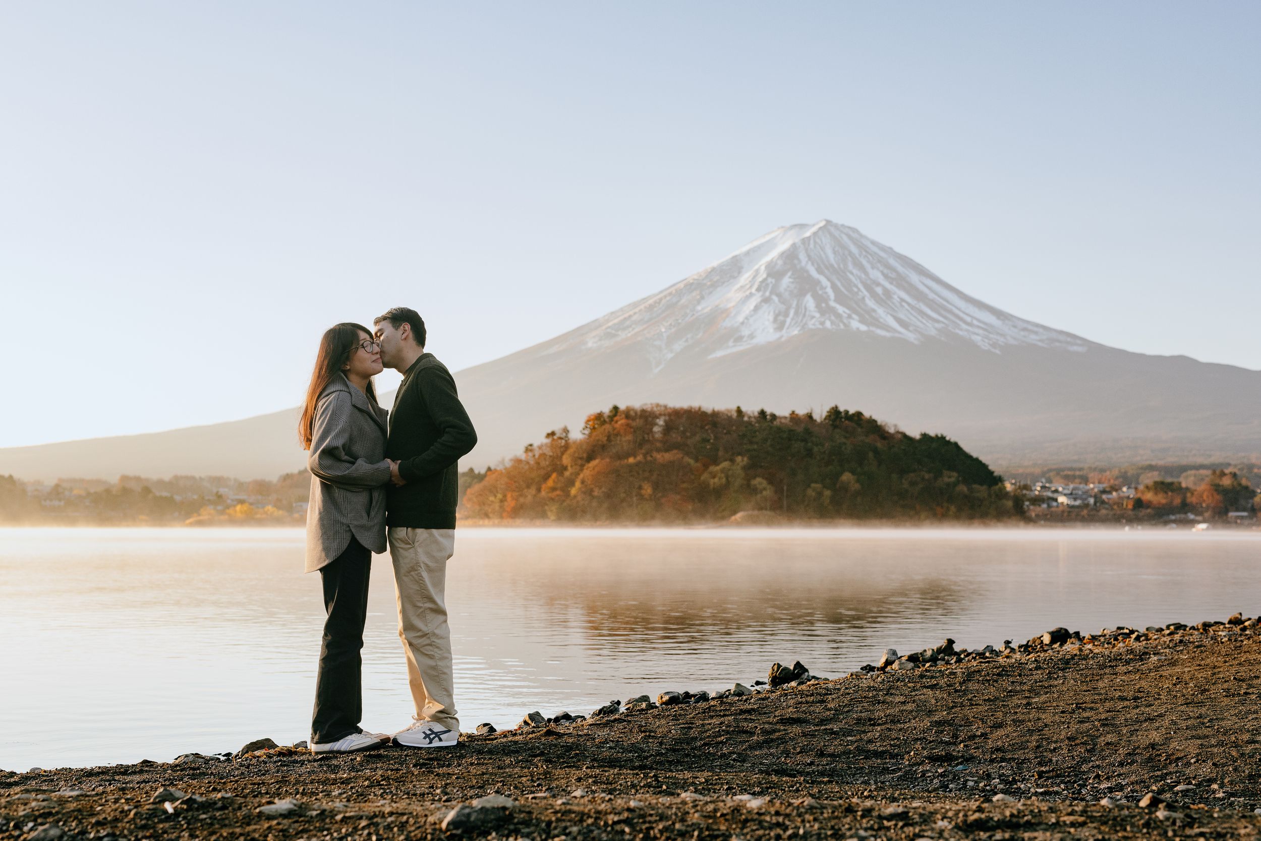 A couple embraces by the shore of Lake Kawaguchiko with Mount Fuji rising majestically in the background.