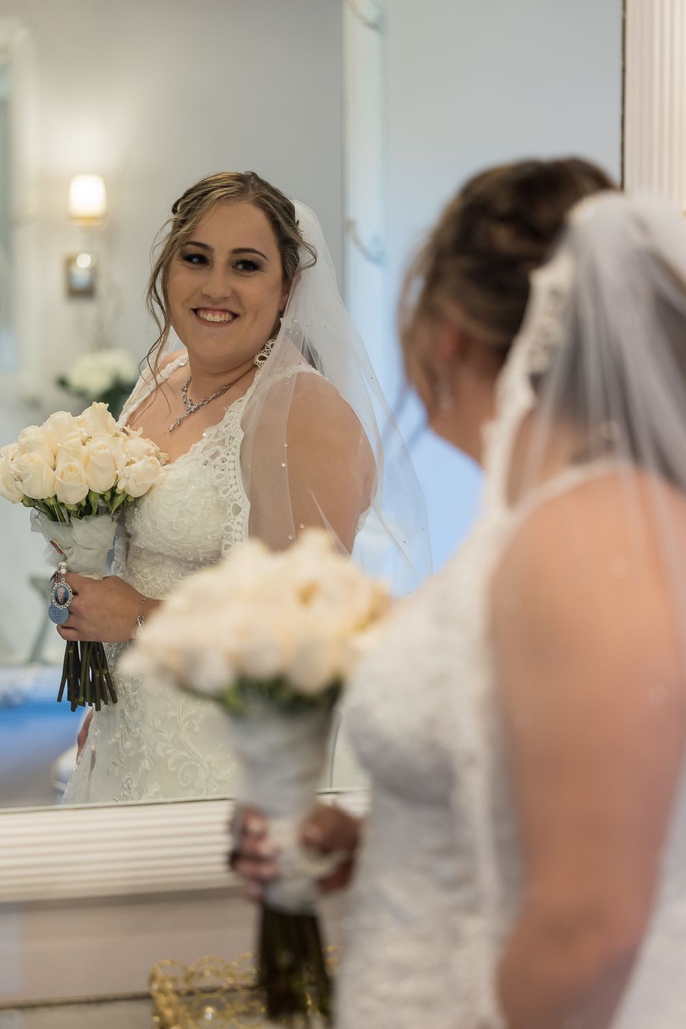 A bride in a white wedding dress holds a bouquet while getting ready in a mirrored dressing room.