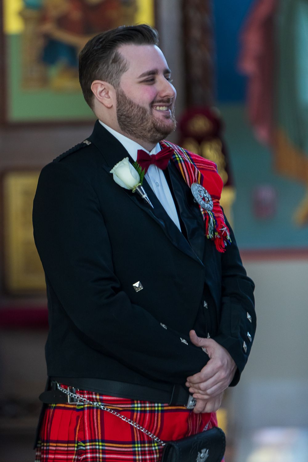 A person in a black suit jacket and red tartan kilt laughs joyfully during what appears to be a formal event.