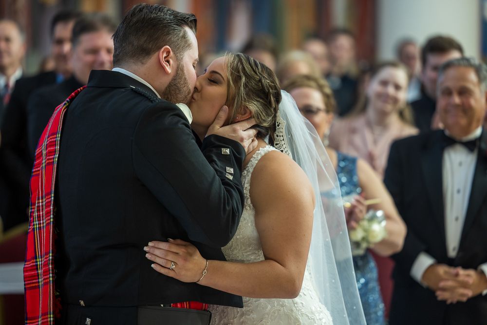 Newlyweds share their first kiss at wedding ceremony while guests look on in background.