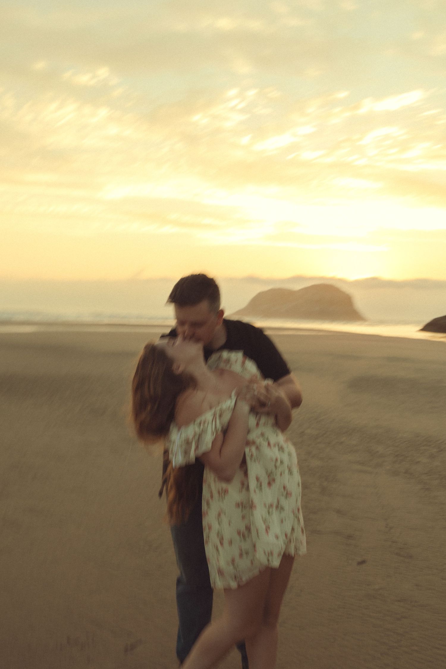 A couple embraces romantically on a sandy beach at sunset with mountains visible in the distance.