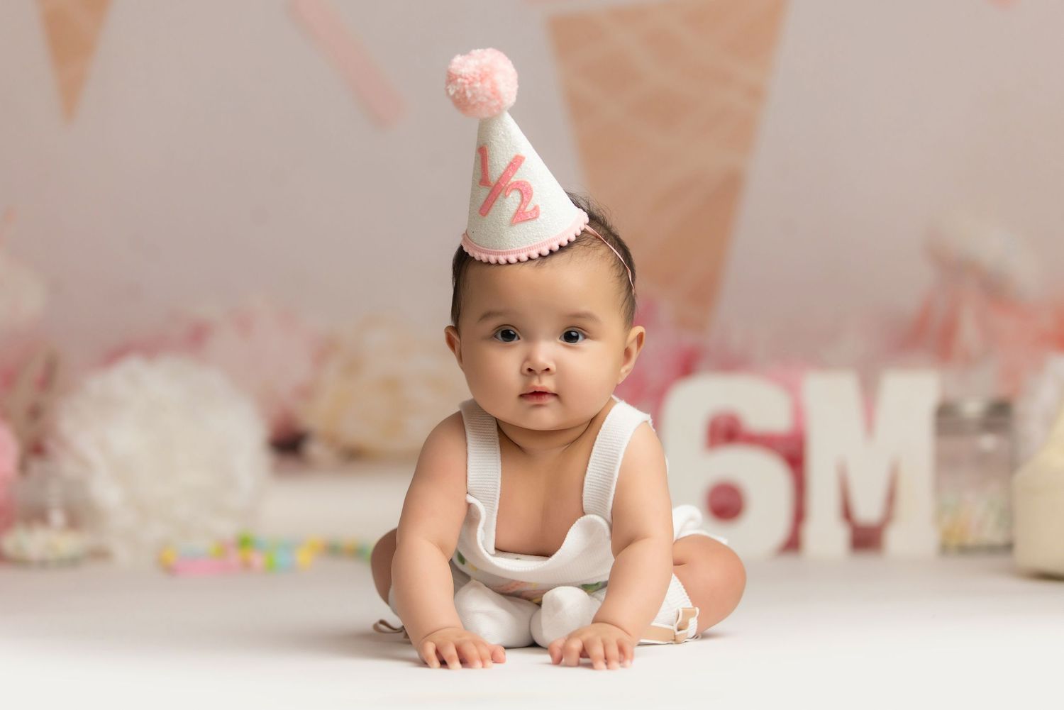 Baby in pink birthday outfit crawls on white backdrop with blush colored decorations in background.