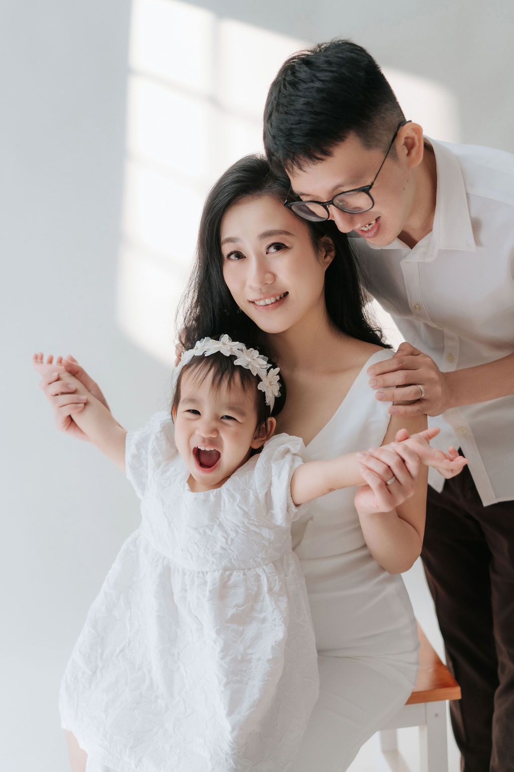 Happy Asian family portrait with smiling mother, father in glasses, and excited toddler girl in white dresses幸福的亞洲家庭合影，照片中笑容燦爛的母親、戴著眼鏡的父親和穿著