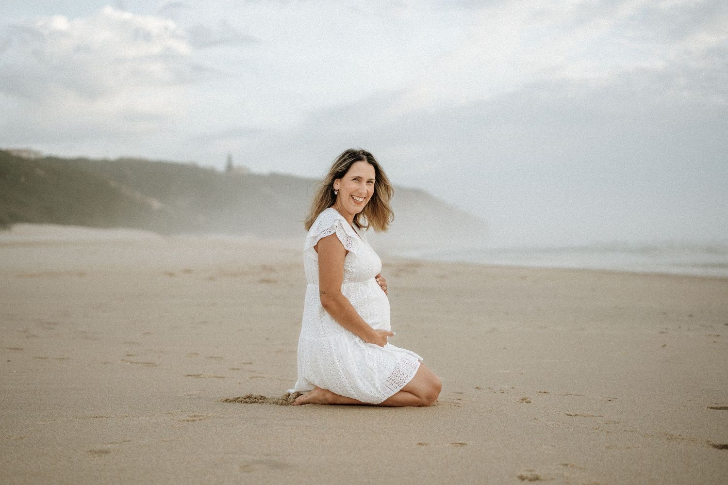 A woman in a white dress twirls gracefully on a misty beach during an overcast day.
