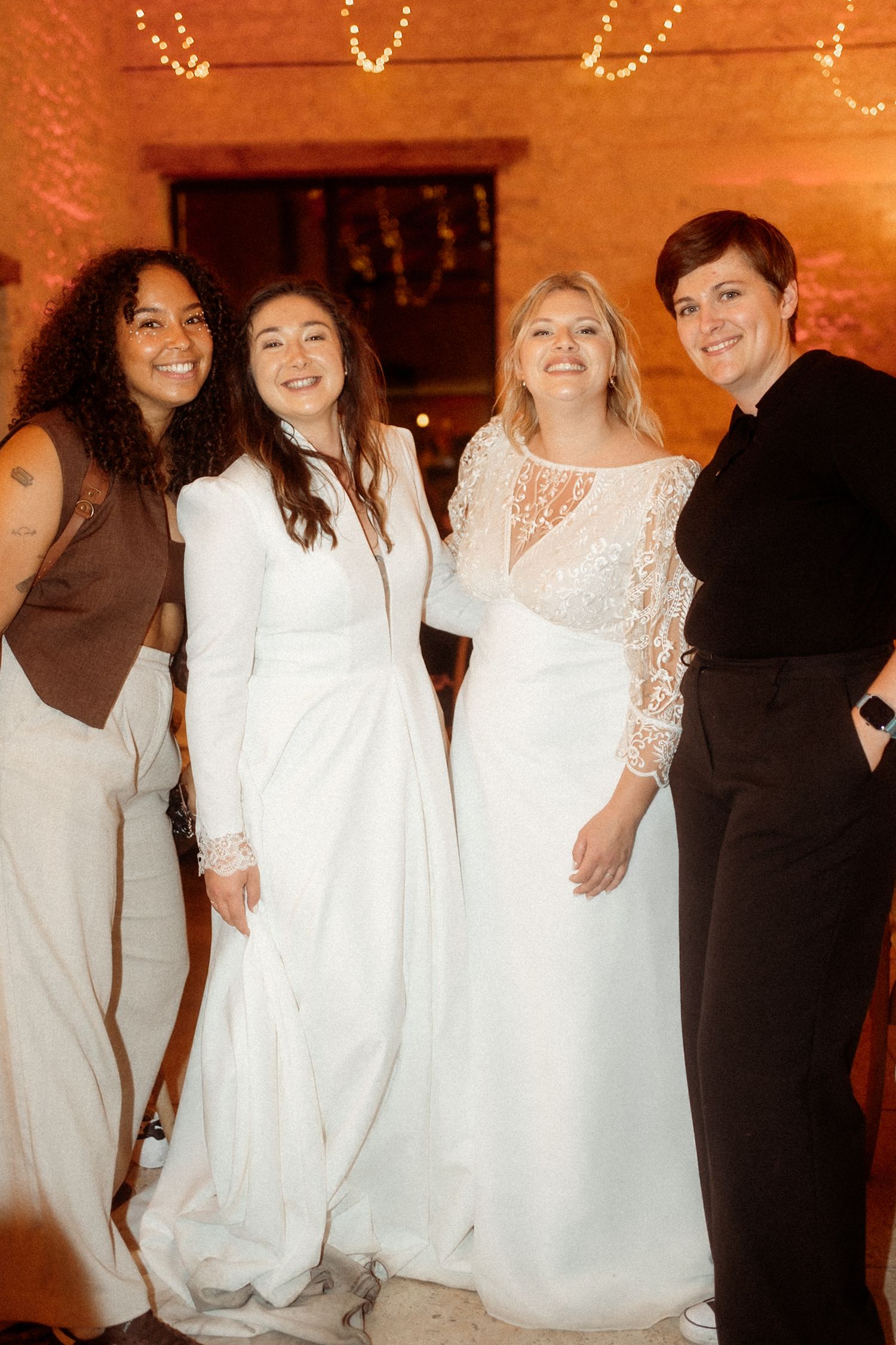 Four friends pose together at a wedding celebration, with three wearing white dresses and one in a brown outfit.