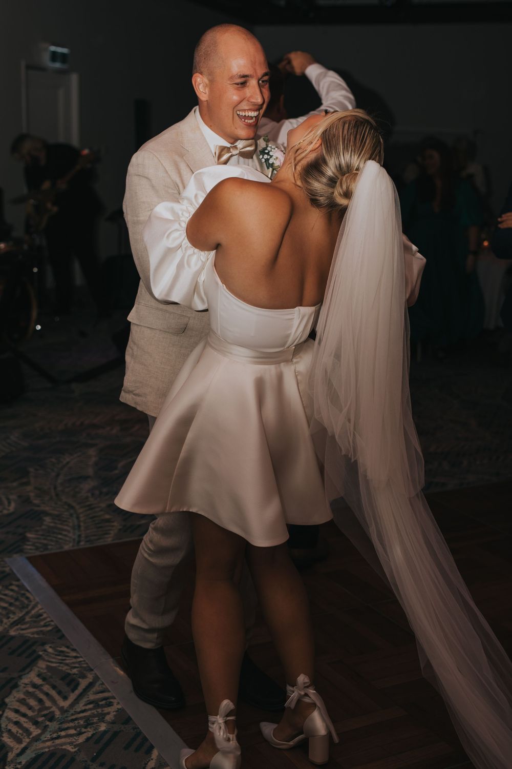 A couple dances together at their wedding reception, with the bride wearing a short white dress with flowing details.