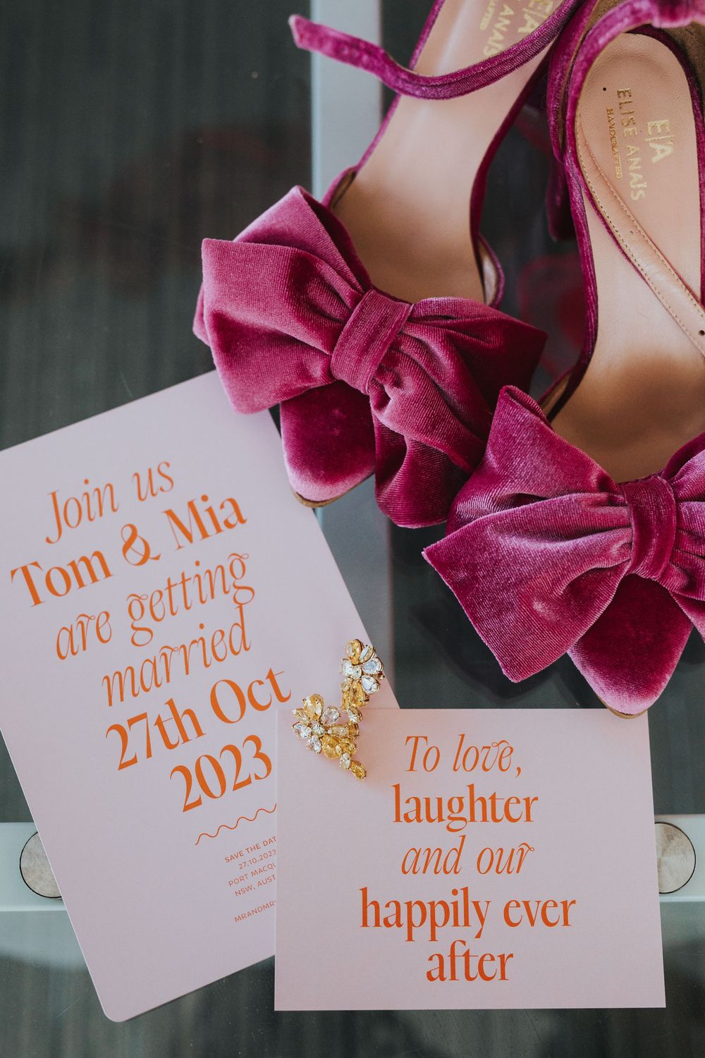 Wedding invitation details in orange calligraphy with pink velvet bow shoes in the background.