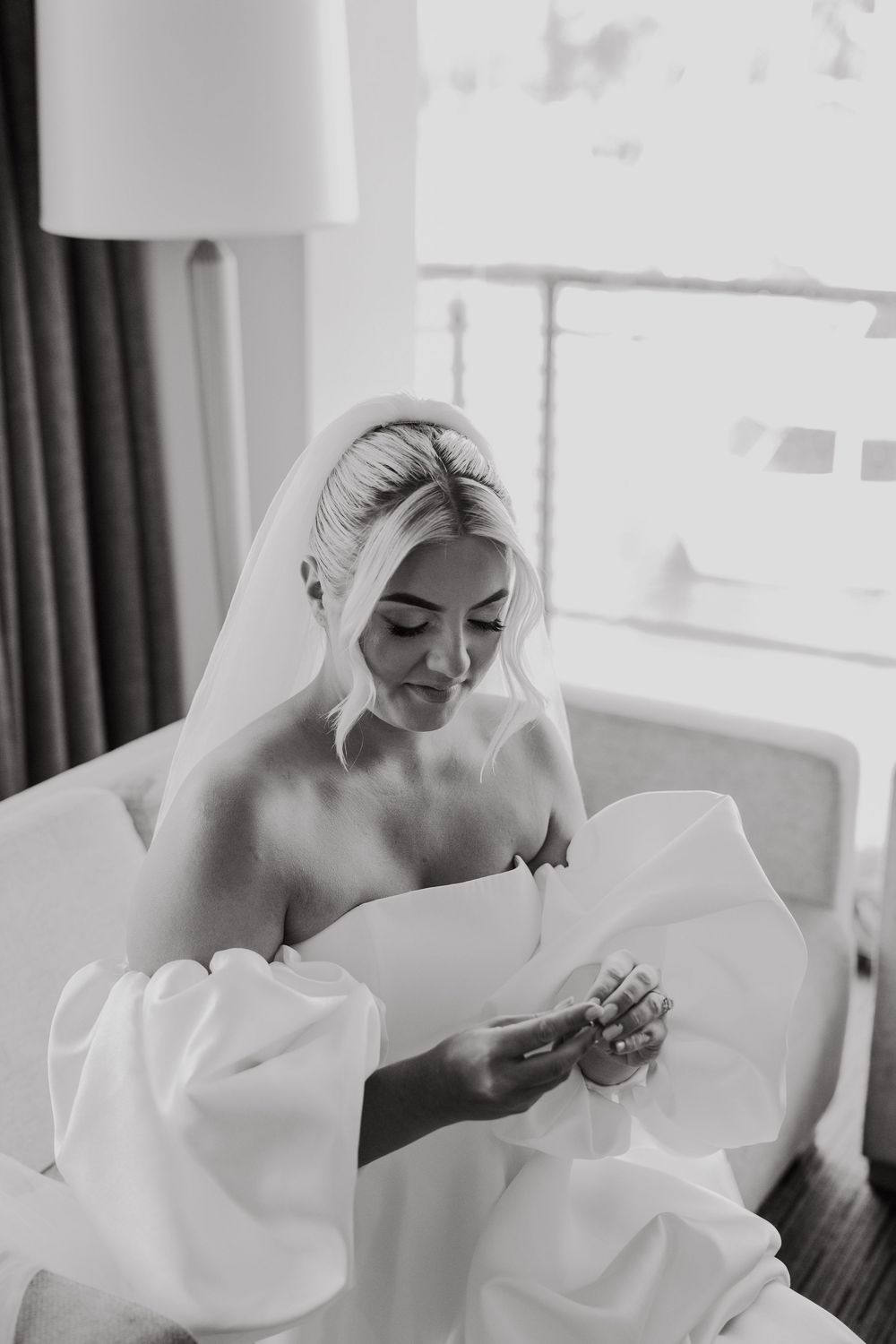 A bride in a white wedding dress adjusts her veil while sitting in a room with natural lighting.