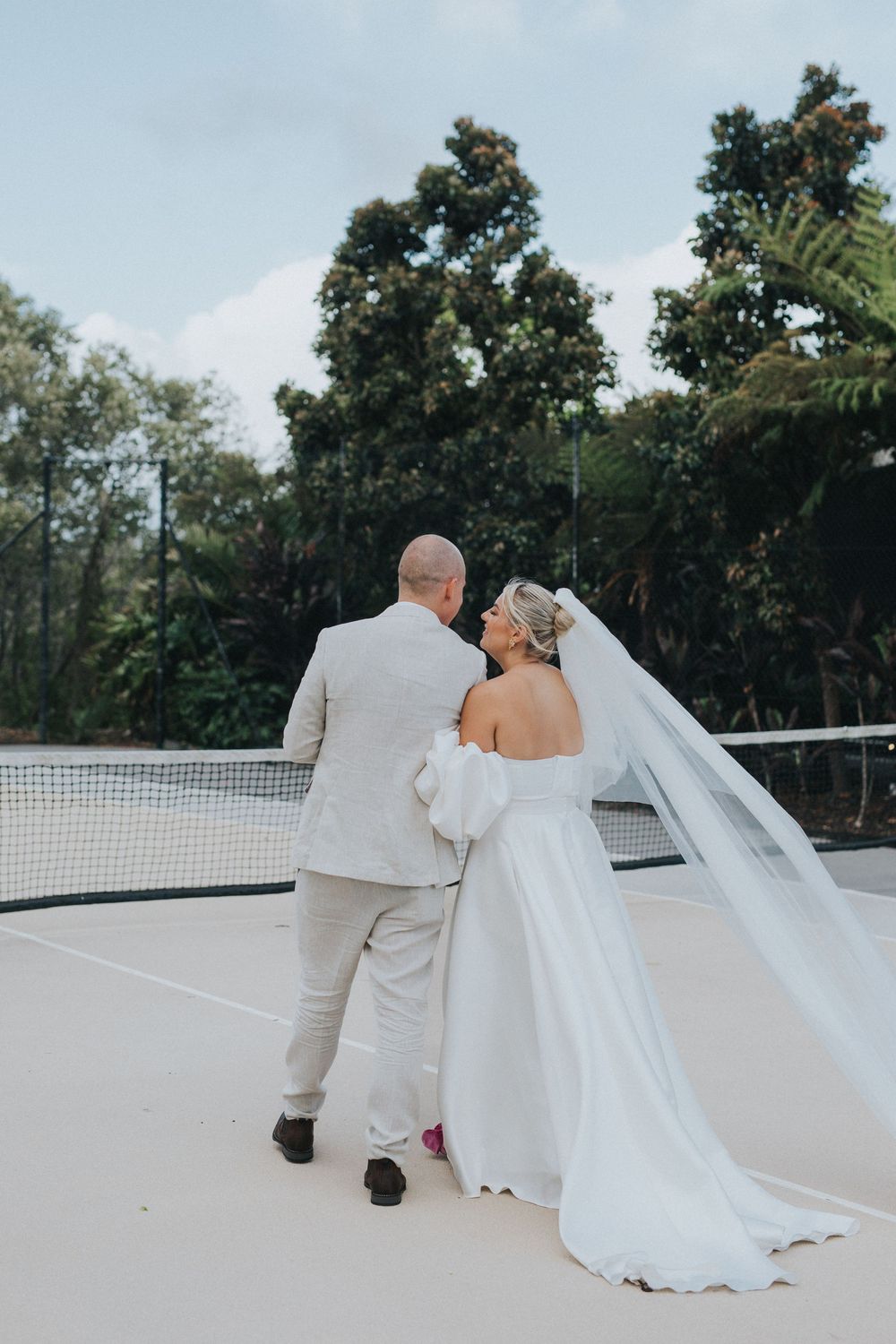 A couple in wedding attire walks along a white balcony with trees in the background.