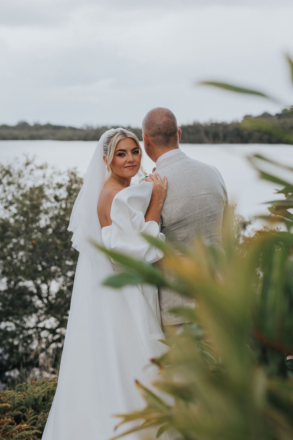 A romantic wedding photo shoot by the water with soft natural lighting and greenery in the foreground.