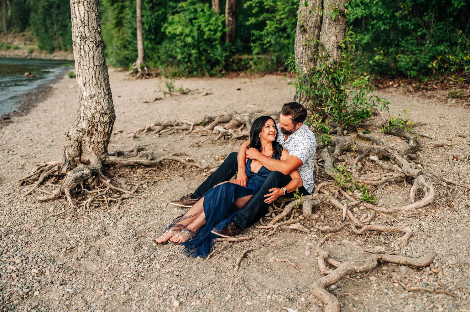 A couple sits together on a sandy beach near tree roots during a romantic sunset photoshoot.