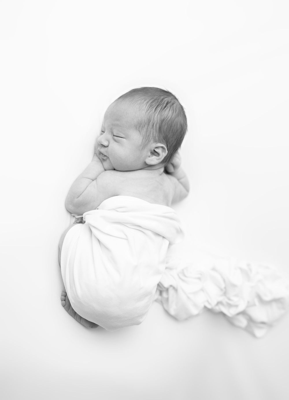 A black and white photo series showing a sleeping newborn baby in white clothing against a white background.