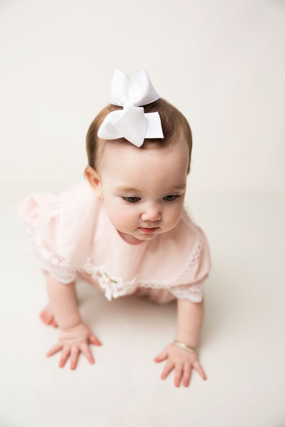 Baby in pink dress and white bow crawls on white background, showing sweet expression.