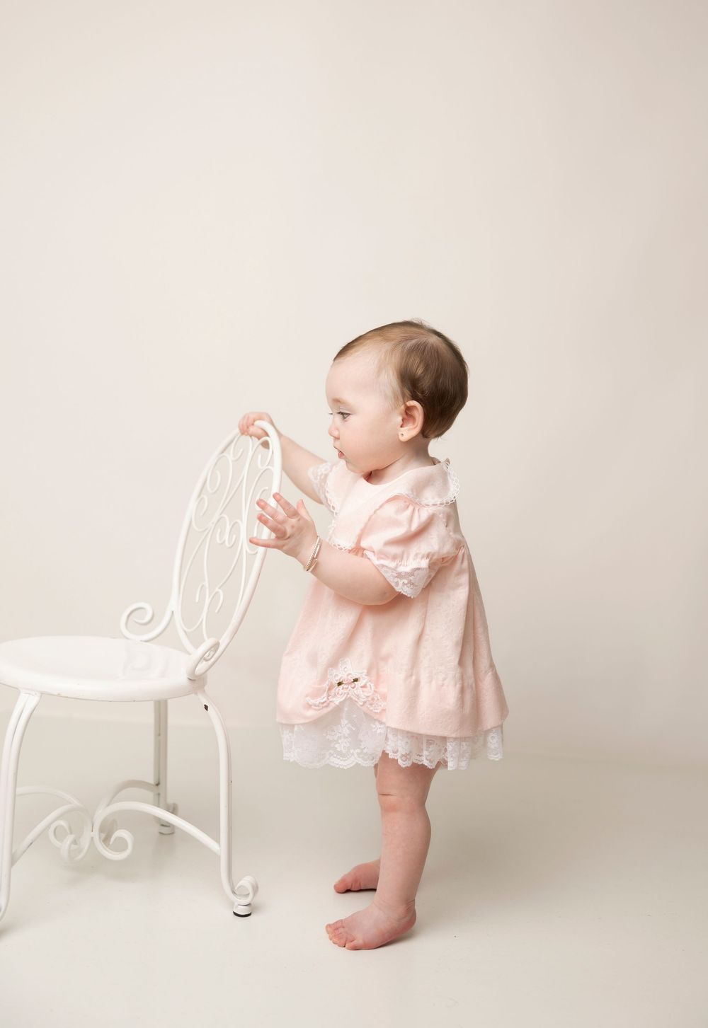 A baby in a delicate pink lace dress explores a white vintage chair against a neutral studio backdrop.