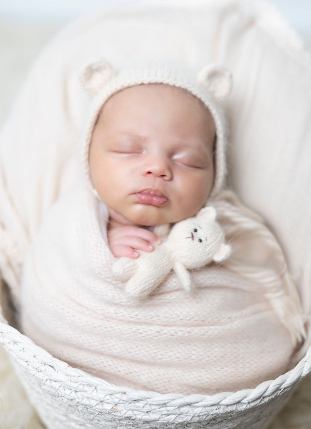 Sleeping baby wearing white knit hat and swaddle blanket holds tiny teddy bear while resting in basket.