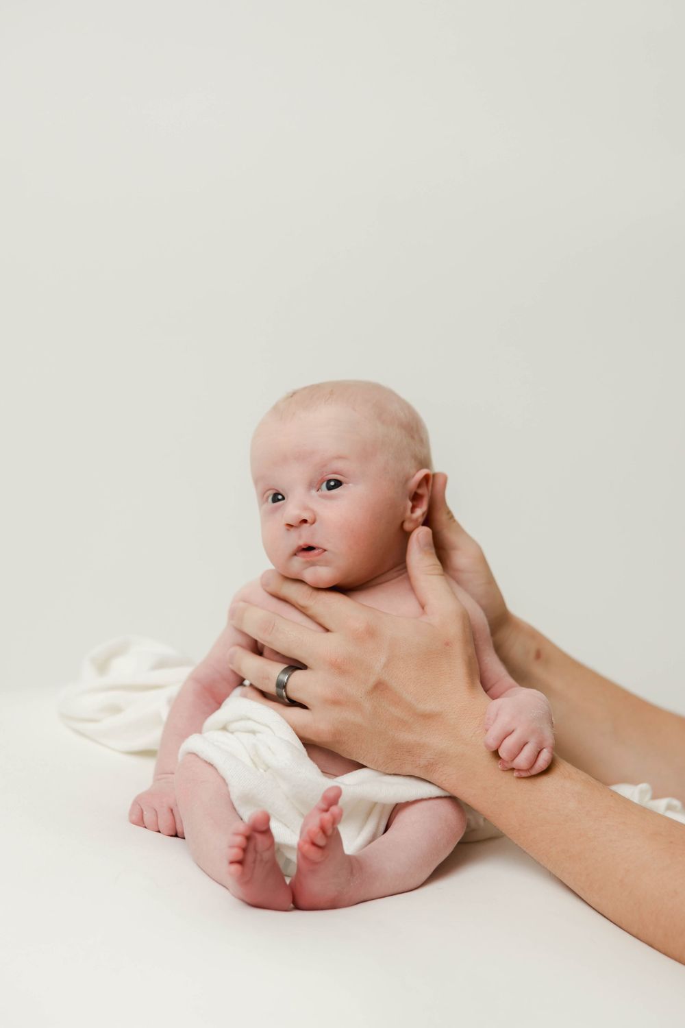 A newborn baby lying on white fabric while gentle hands provide support during a professional photo session.