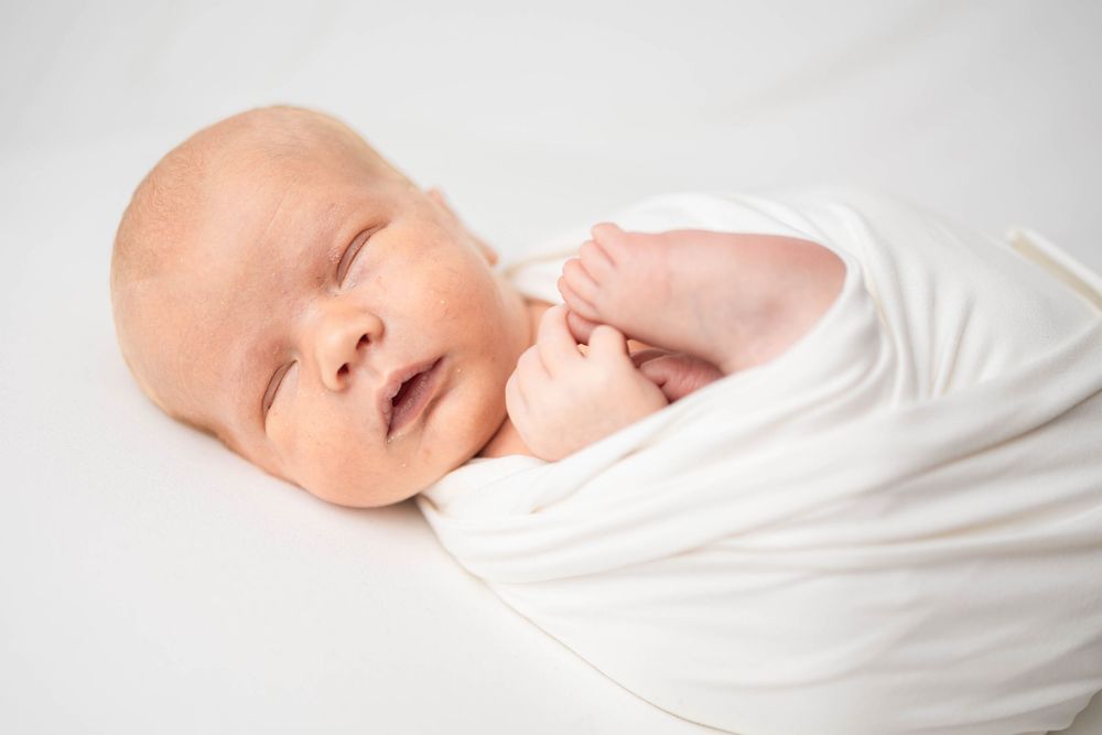 A peaceful sleeping newborn wrapped in soft white fabric against a light background.