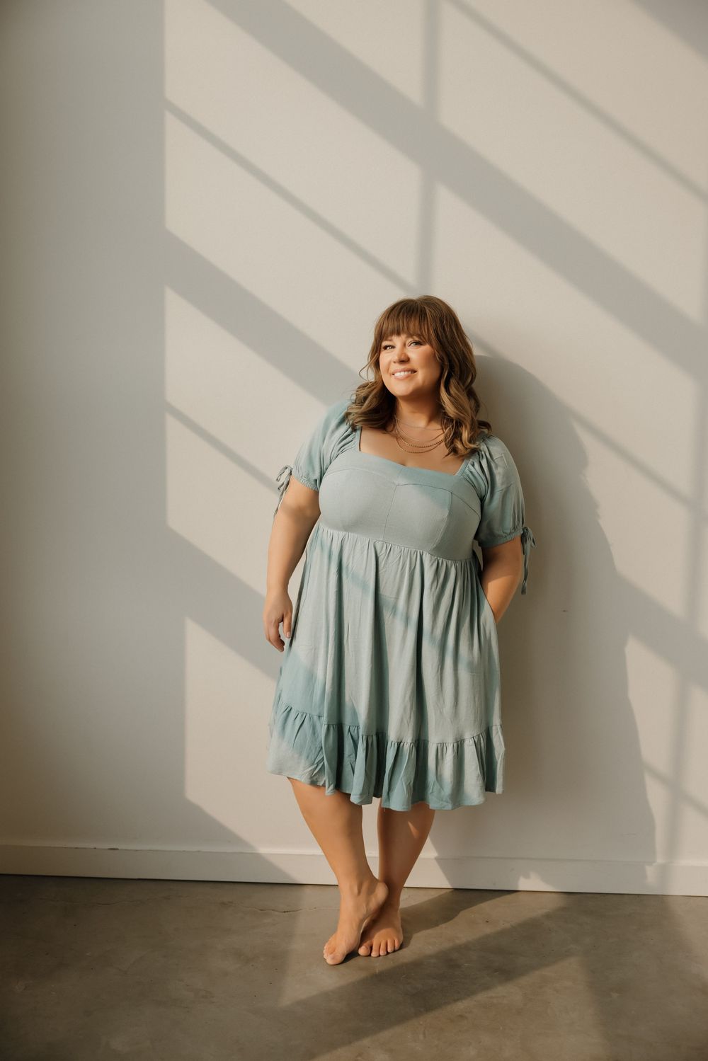 Woman in sage green tiered ruffle mini dress with puff sleeves, posing barefoot against a white wall.