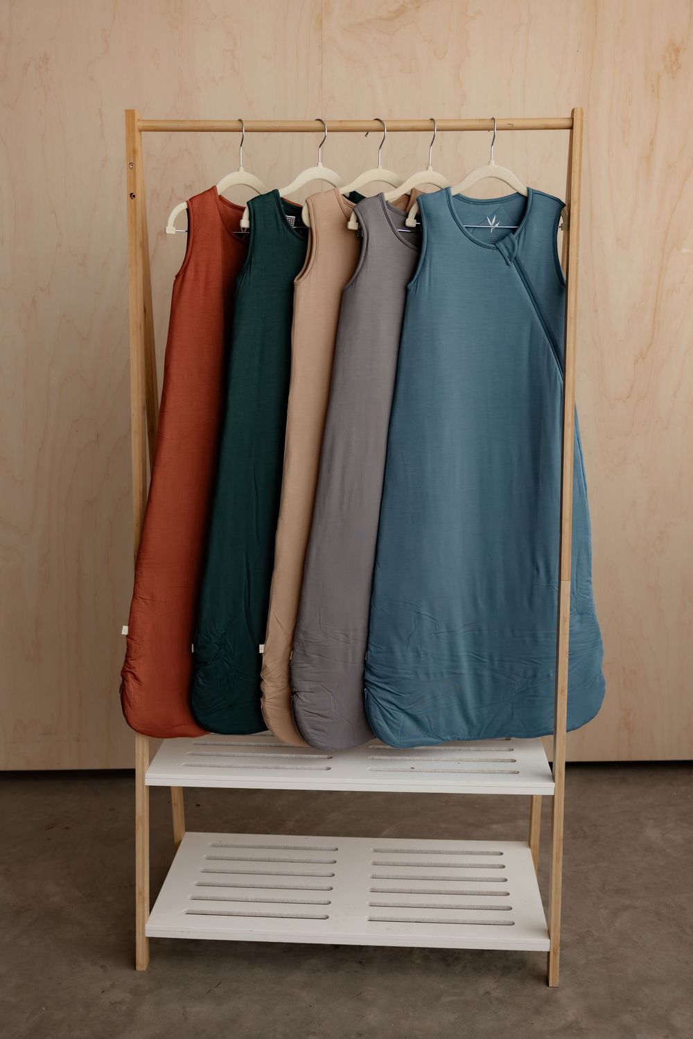 Five colorful sleeveless baby sleep sacks hanging on a wooden clothing rack in rust, green, tan, gray, and teal.