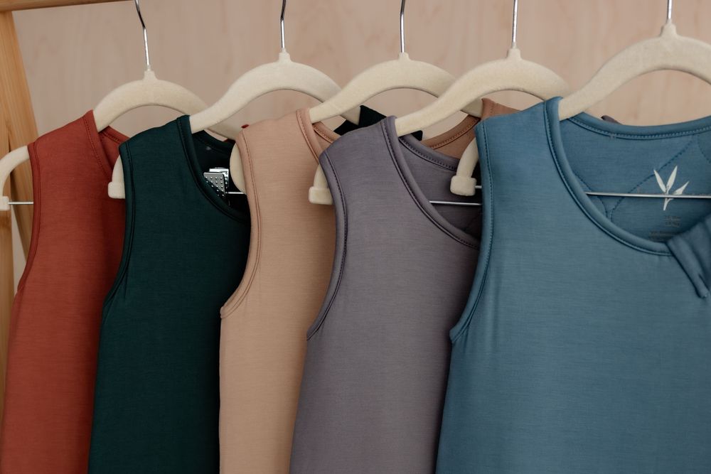 Colorful sleeveless tank tops in rust, teal, beige, gray, and blue hanging on wooden hangers.