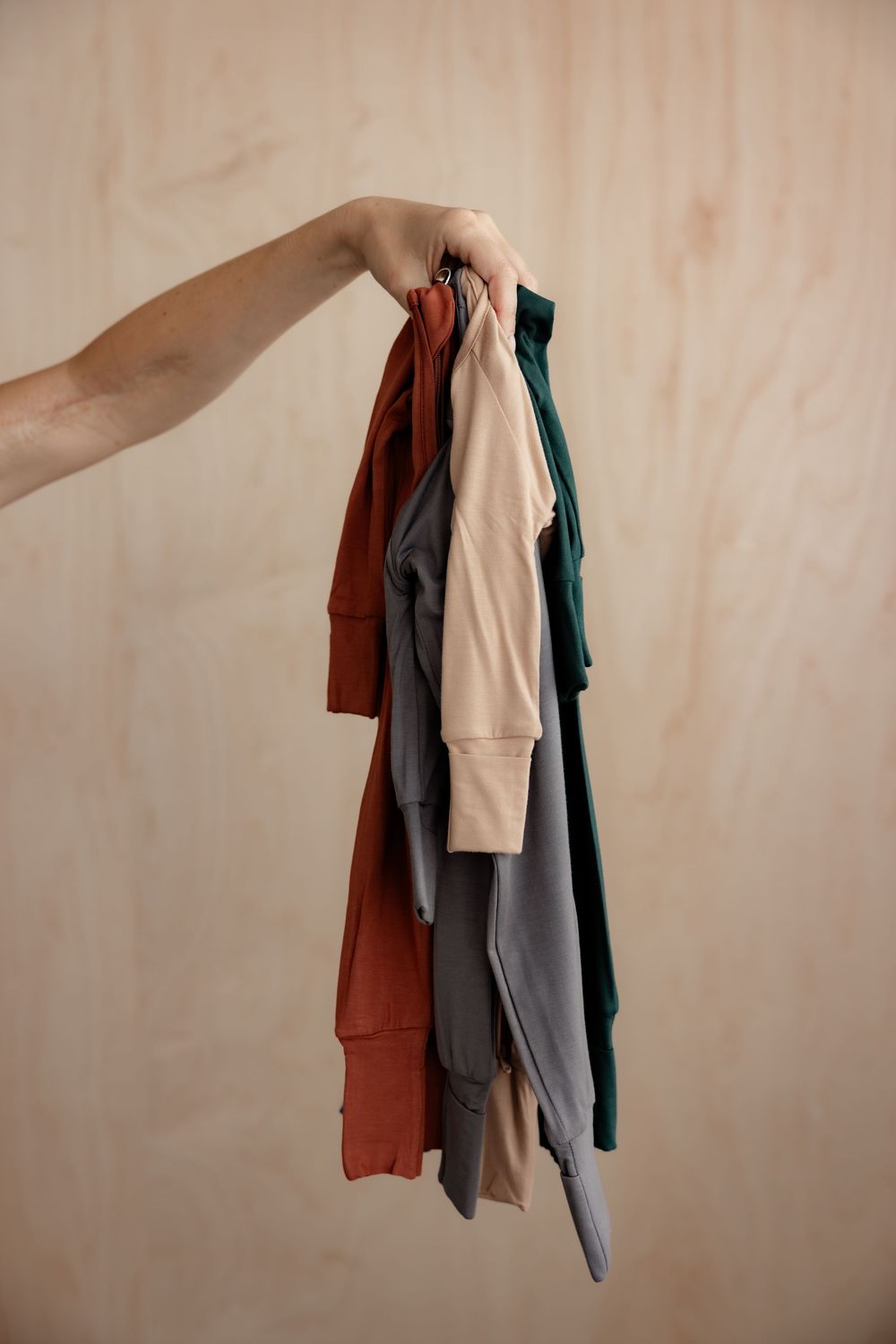 Hand holding multiple folded t-shirts in rust, beige, gray, and green colors against a light wood background.