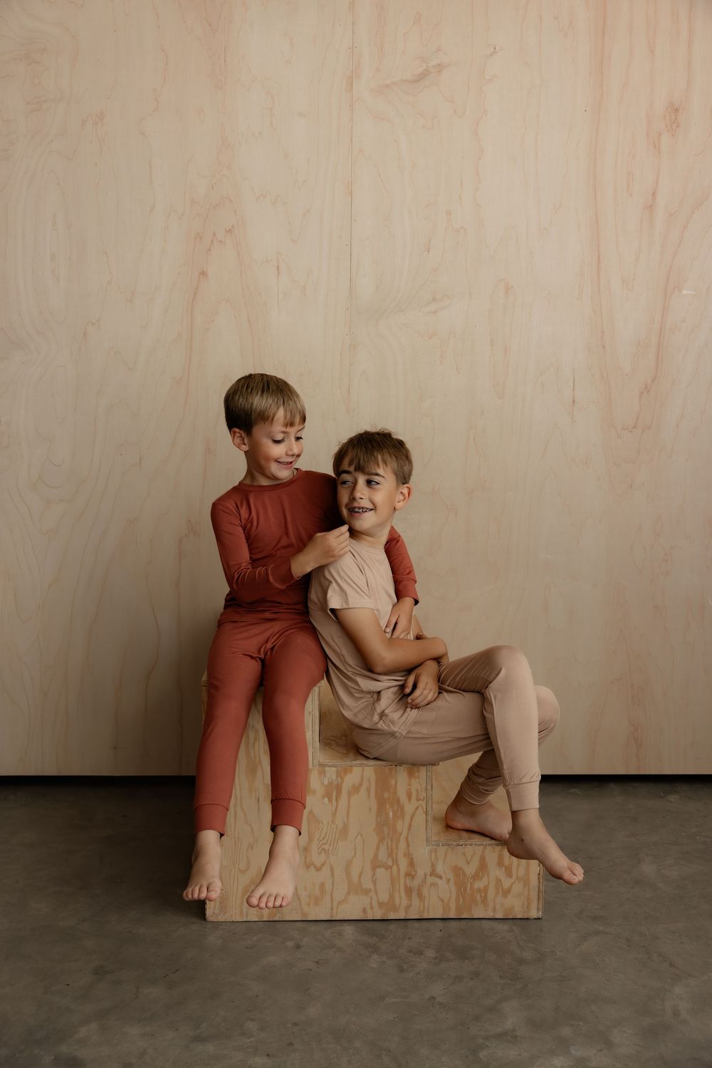 Two boys in matching earth-tone loungewear sitting on a wooden box against a plywood wall.