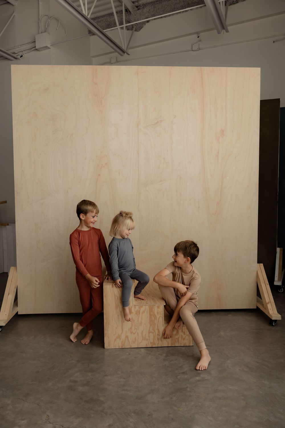 Three barefoot children in colorful loungewear playing on a wooden cube against a plywood backdrop in a studio.