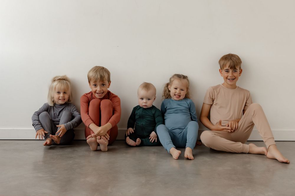Five young children in colorful matching pajamas sitting together against a white wall on a gray floor.