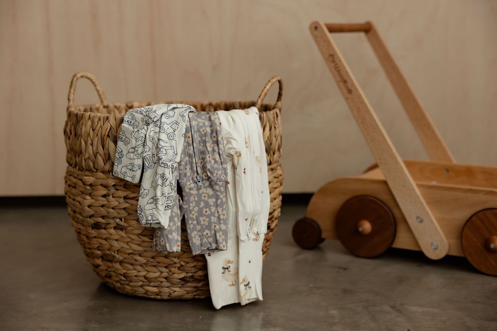 Wicker basket with floral baby clothes beside a wooden push toy cart on a gray floor.
