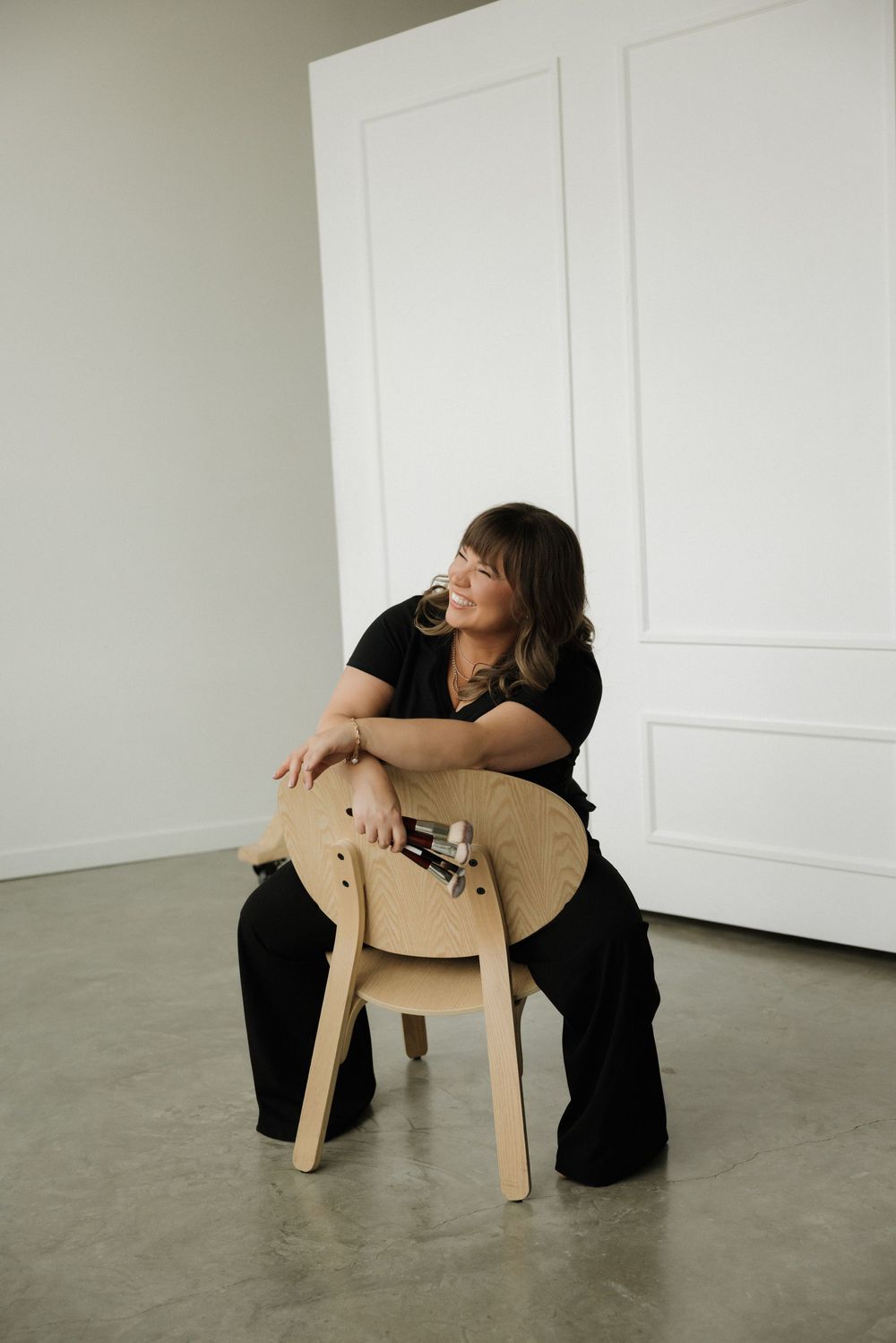 Woman in black outfit sitting backwards on a wooden chair in a bright white studio with concrete floors.