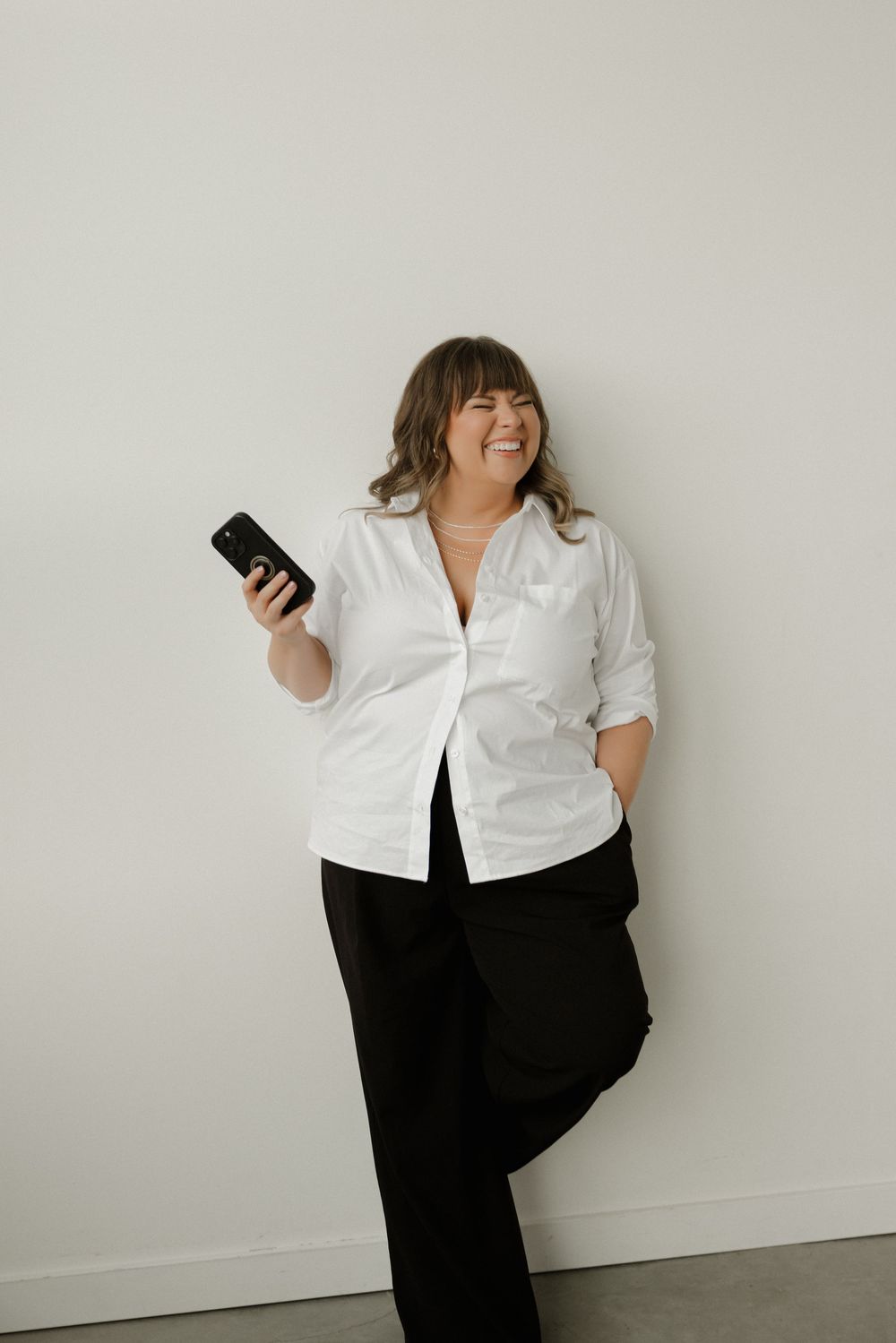 Woman in white shirt and black pants leaning against wall holding smartphone, smiling confidently.