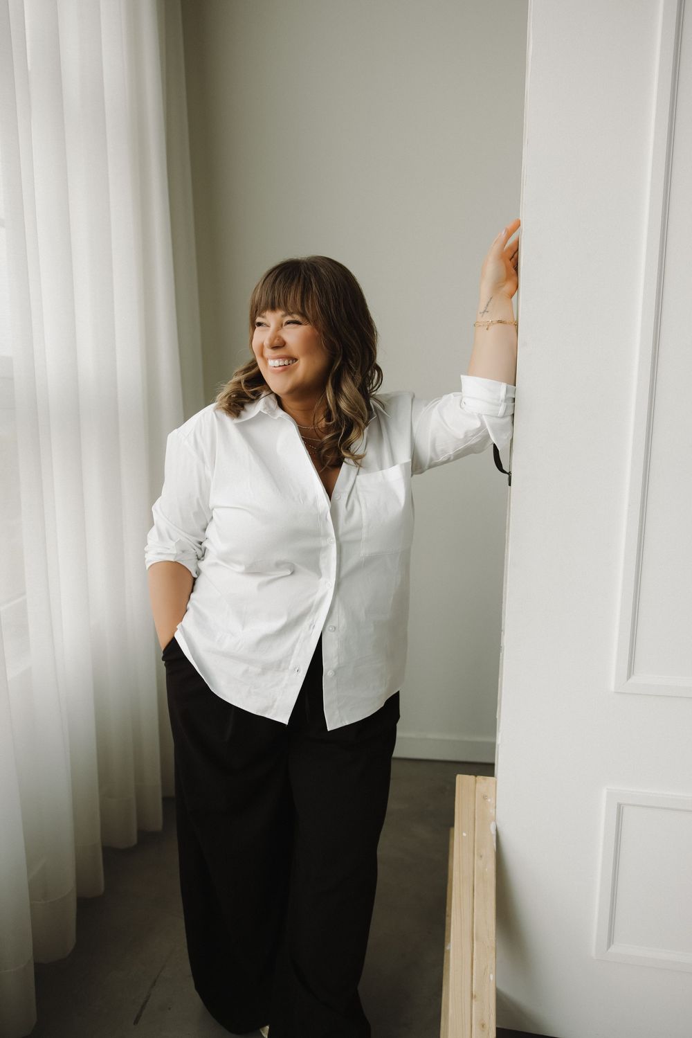 Woman in white button-up shirt and black pants leaning against a white door frame, smiling softly.