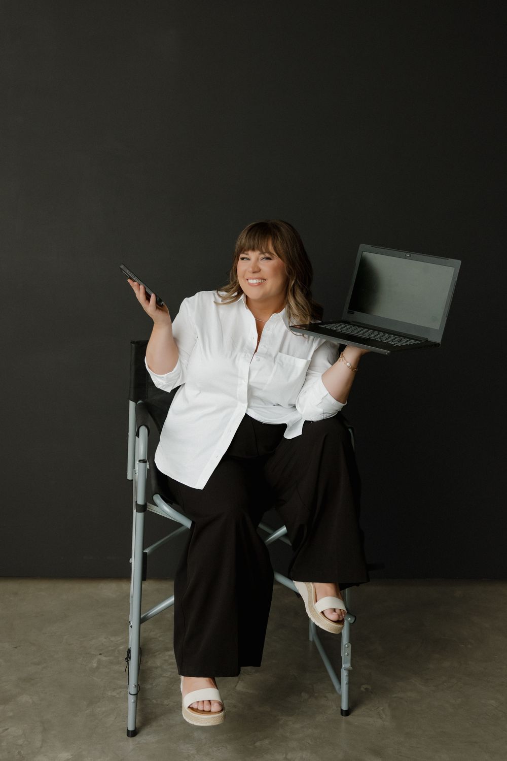 Woman in white shirt holding laptop and phone while sitting on director's chair against dark background.