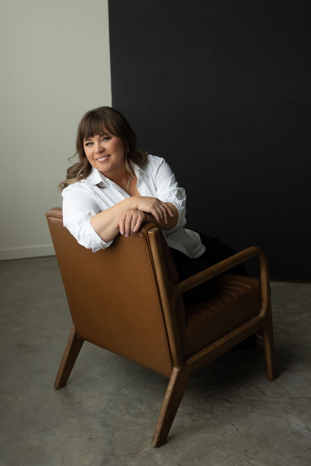 Woman in white shirt sitting relaxed in a mid-century modern brown leather chair against a dark wall.