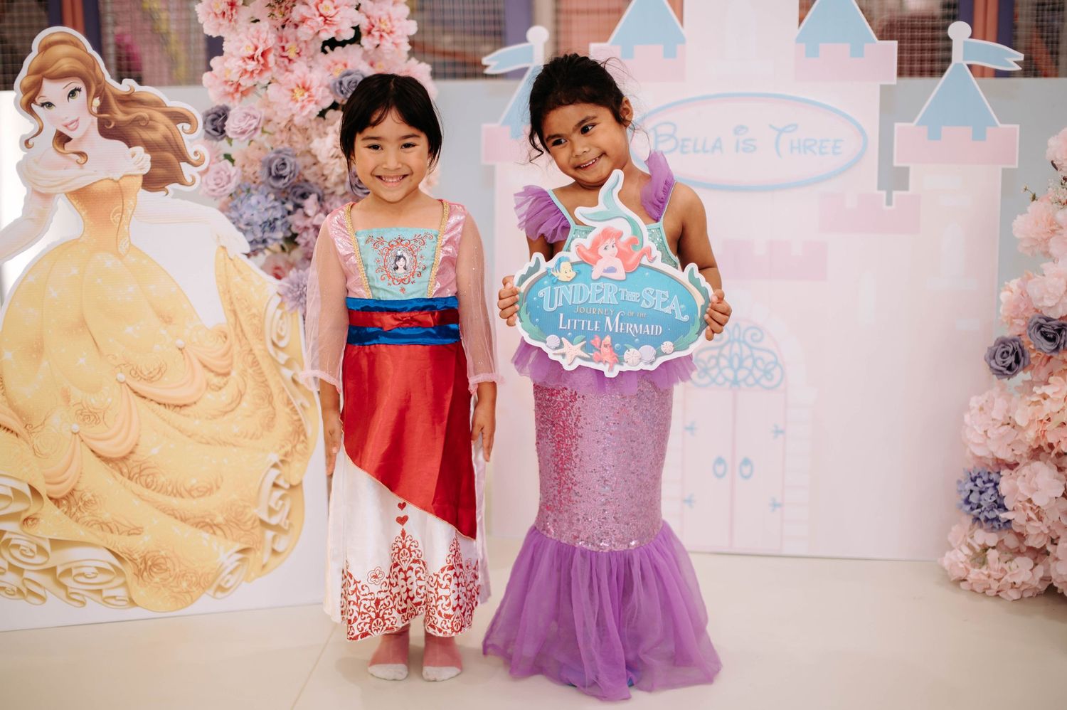 Two young children wearing princess costumes pose in front of Disney princess cutouts and pink decorations.