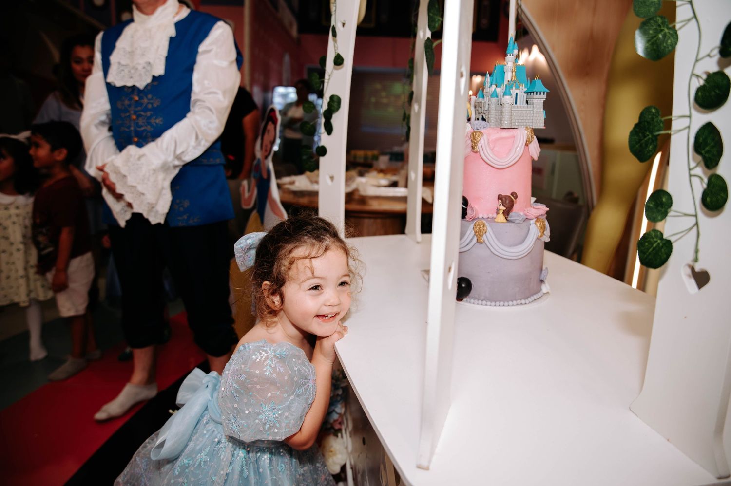 A child in a blue dress poses near a princess-themed birthday cake and decorative elements.