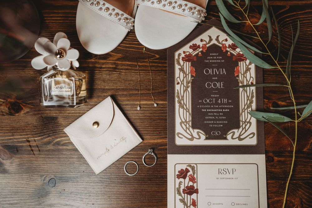 Wedding details flatlay with invitation suite, rings, perfume and bridal shoes on wooden surface.