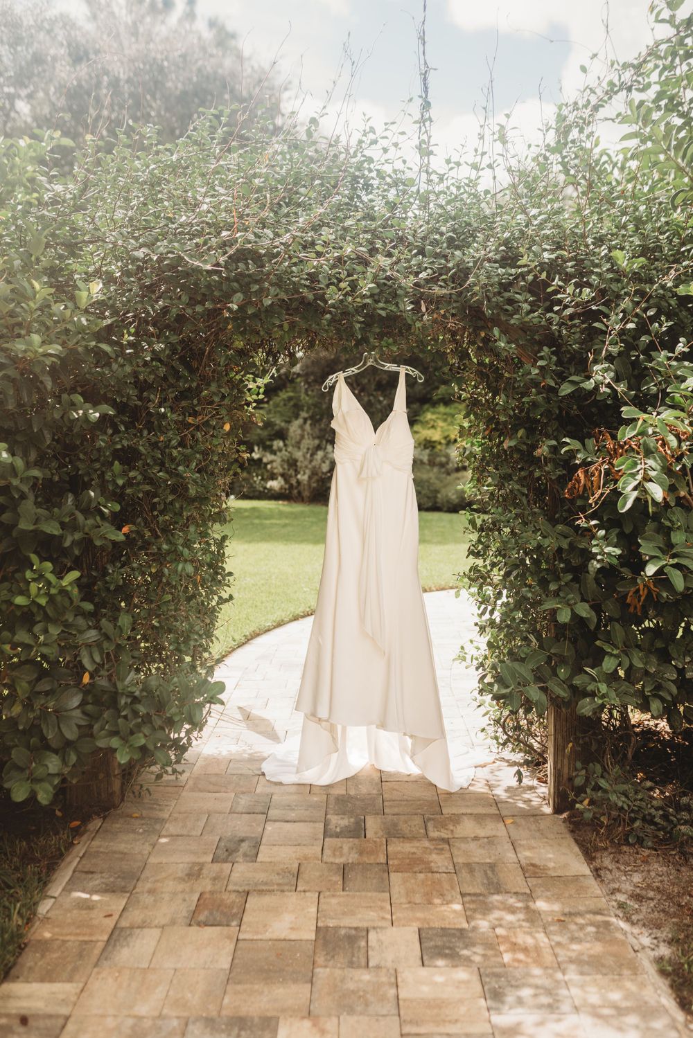 An elegant white wedding dress hangs in a garden archway with stone pathways.